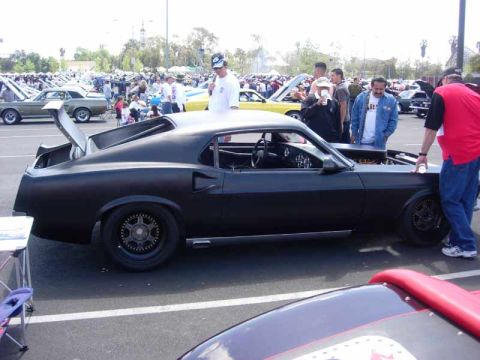 1998 Ford Crown Victoria - matte-black fastback muscle car lowered on black wheels with a tall rear wing sits in profile as a crowd mills around it.