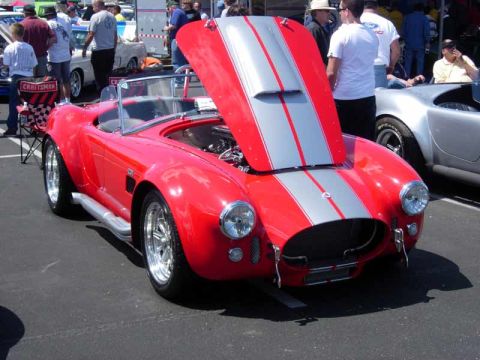 1998 Ford Crown Victoria - red Cobra-style roadster with twin silver stripes sits with its hood raised, parked beside a bare-aluminum Cobra at a show.