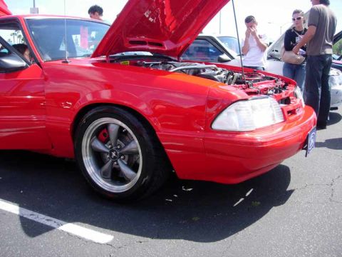 1998 Ford Crown Victoria - red Fox-body Mustang with its hood open and gray five-spoke wheels is shown from the front corner as spectators stand nearby.