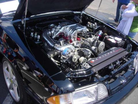 1998 Ford Crown Victoria - open engine bay of a black car reveals a polished supercharged motor with a valve cover labeled Chicane.