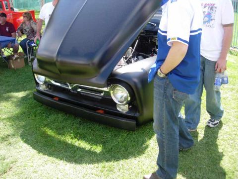 1998 Ford Crown Victoria - matte-black 1950s Ford pickup sits hood-up on grass with chrome bumper and round headlights as people stand alongside.