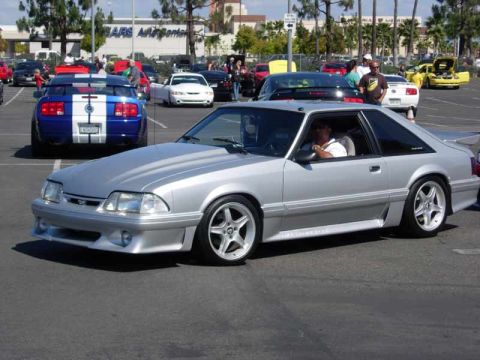 1998 Ford Crown Victoria - silver Fox-body Mustang GT with five-spoke wheels rolls through a car-show lot past a blue Shelby and other Mustangs.
