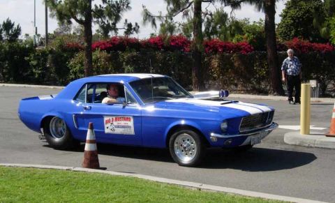 1998 Ford Crown Victoria - blue first-gen Mustang coupe drag car with white stripe, hood scoop and door lettering turns a corner past a traffic cone.