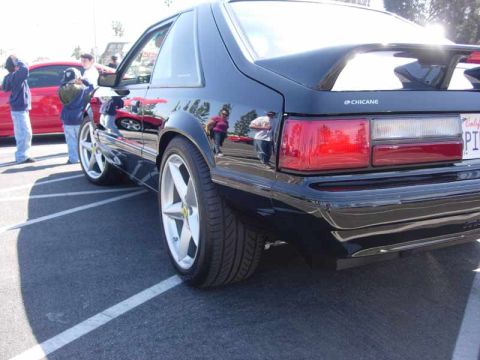 1998 Ford Crown Victoria - rear corner of a glossy black Fox-body Mustang shows Chicane badging and silver Ferrari-emblem wheels in a parking lot.