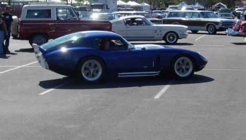 1998 Ford Crown Victoria - blue Shelby Daytona Coupe replica sits in profile in a parking lot, with a red truck and a white Mustang fastback behind.