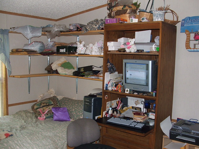 Cluttered bedroom with shelves full of items, a bed, a desk with a computer monitor, and a chair