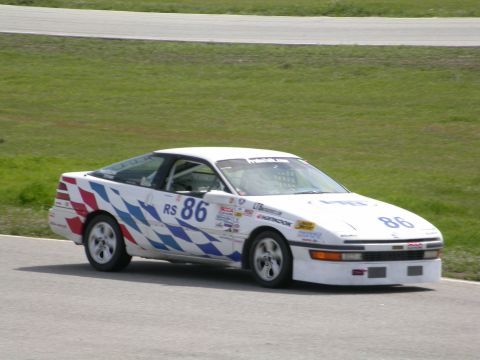1990 Ford Probe - A white race car with blue and red checkered graphics and the number 86 is seen on a race track.