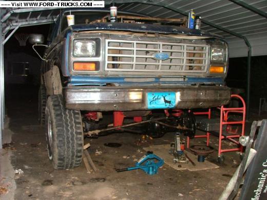 Front view of a blue Ford truck with its front end raised on jacks, undergoing repairs. Tools and parts are scattered around
