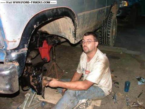 1985 Ford Bronco - A mechanic works on the front suspension of a muddy SUV, with another vehicle in the background.