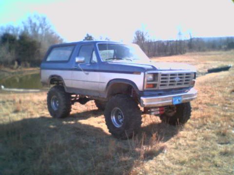 1985 Ford Bronco - This lifted Ford Bronco is parked in a dry, grassy field with trees and a pond in the background.