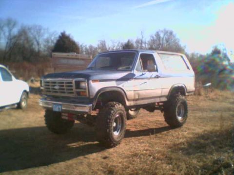 1985 Ford Bronco - A lifted Ford Bronco sits in a grassy field under a bright blue sky, with another vehicle to its left.