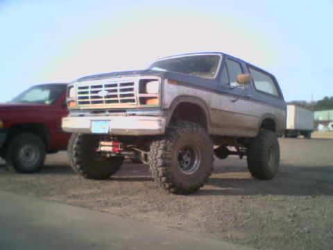 1985 Ford Bronco - This lifted Ford Bronco is parked next to a red pickup truck under a clear sky.