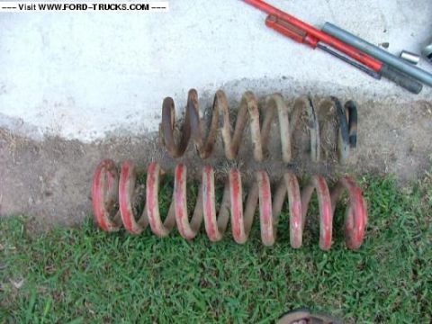 1985 Ford Bronco - Two sets of coil springs, one painted red and the other showing rust, are displayed outdoors on grass and dirt.