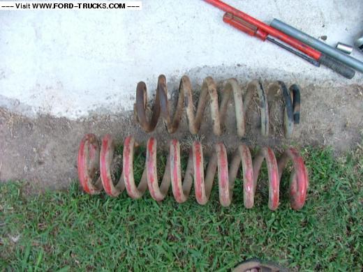 Two sets of used coil springs, one red and one rusty brown, lie on grass and dirt