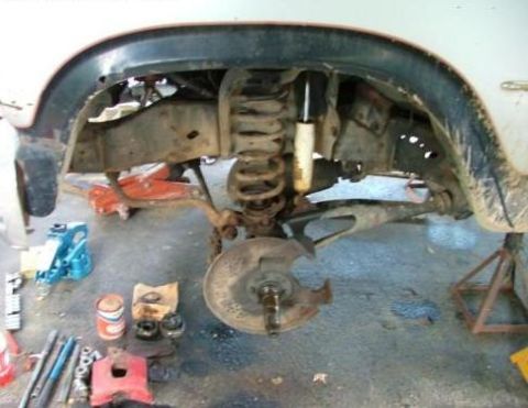 1985 Ford Bronco - The front suspension of a vehicle is disassembled and supported by jack stands, with various tools and parts visible on the ground.