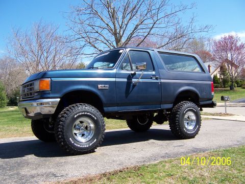 1987 Ford Bronco - A lifted blue Ford Bronco is shown from the side on a sunny day with trees in the background.
