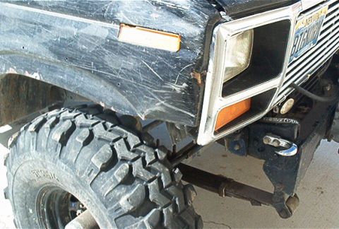 1986 Ford Bronco - The front corner of a heavily scratched black Ford Bronco is shown, with a large off-road tire and suspension components visible.
