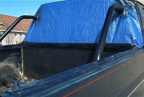 1986 Ford Bronco - Roll bar and bedliner covered by a blue tarp, with a house in the background.