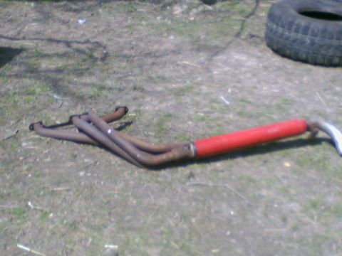 1978 Ford Bronco - A rusty exhaust manifold with a red muffler pipe lies on the ground next to a tire.