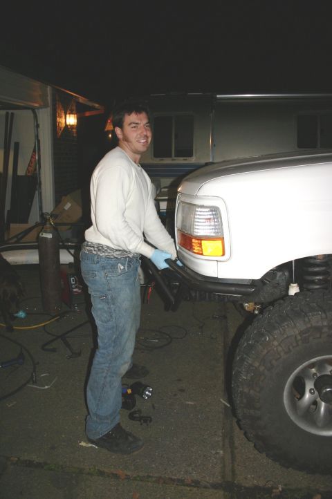 1995 Ford Bronco - A man works on the front bumper of a white SUV, which appears to be from the early to mid-1990s.