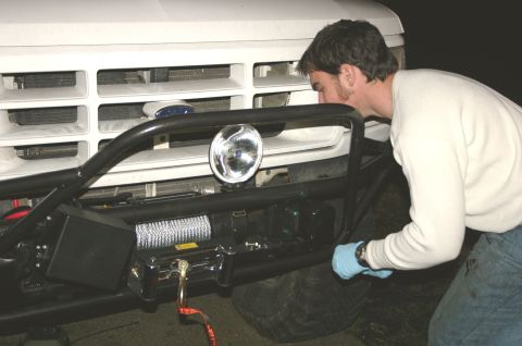 1995 Ford Bronco - A man works on the front bumper of a white 1995 Ford Bronco, which is equipped with a winch and a large spotlight.