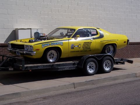 1972 Ford Ranchero - A modified yellow car with a prominent engine sits on a trailer outside a brick building.