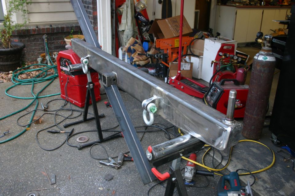 Custom steel bumper with D-ring mounts and a hitch receiver, resting on saw horses with welding equipment nearby