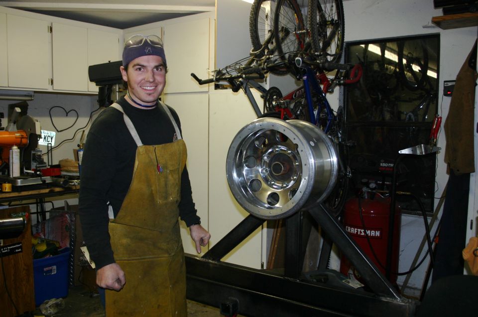 Man in workshop wearing apron and goggles, standing next to a large tire rim mounted on a stand. Bicycles are hanging on the wall behind him