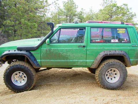 1994 Ford Bronco - A green Jeep Cherokee with a snorkel and off-road tires is parked on a dirt road.
