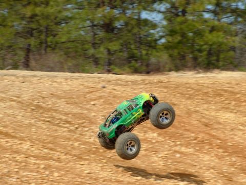 1994 Ford Bronco - RC monster truck mid-air jump on a dirt track with trees in the background.