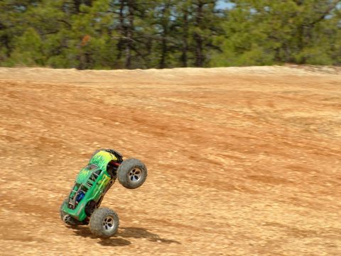 1994 Ford Bronco - A green remote control monster truck is captured mid-air performing a wheelie on a dirt track with trees in the background.