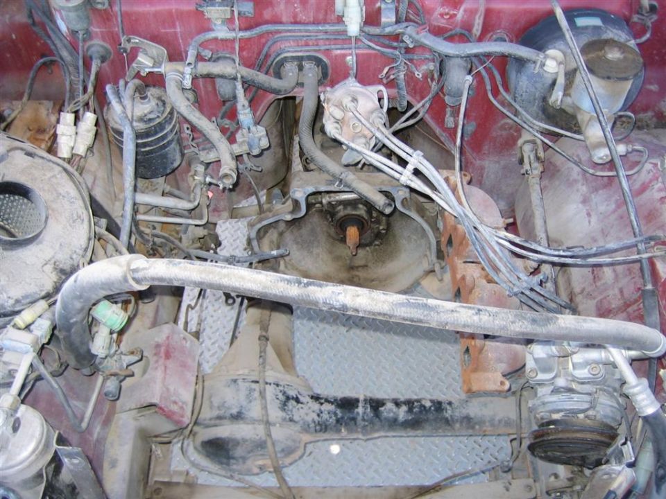 Dusty red 4x4 engine bay from above with distributor, plug wires, rusty manifold and diamond plate