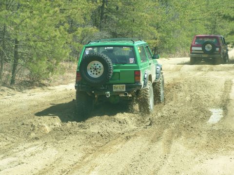 1994 Ford Bronco - Two off-road vehicles navigate a dirt path with mud and trees.