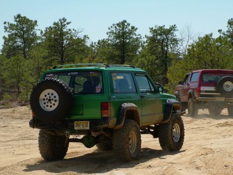 1994 Ford Bronco - A lifted green Jeep Cherokee is seen from the rear, with a red SUV following on a dirt path surrounded by trees.