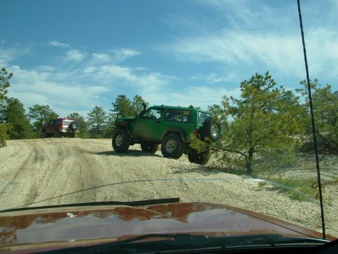 1994 Ford Bronco - Two off-road vehicles navigate a dusty trail with trees and blue sky in the background.
