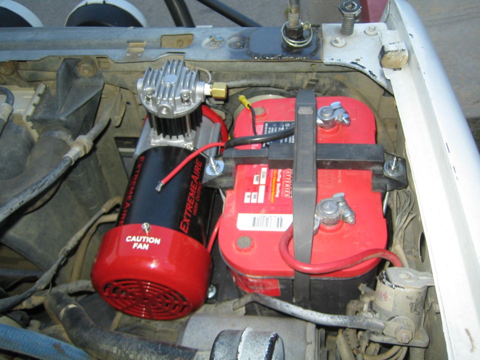 Engine bay with a black ExtremeAire air compressor mounted beside a red battery secured by straps