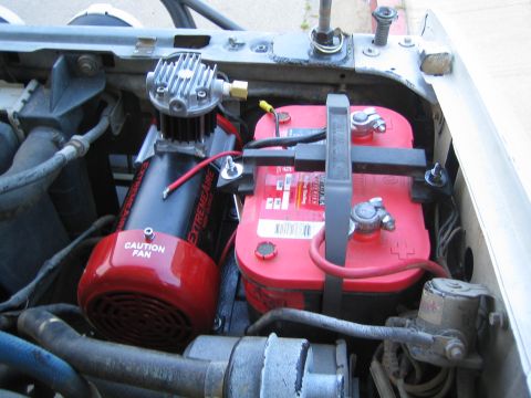 1990 Ford Bronco - Close-up in an engine bay of a black ExtremeAire air compressor mounted beside a red battery held by black straps.