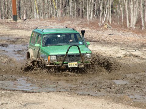 1994 Ford Bronco - A green Jeep Cherokee navigates a muddy off-road course, kicking up a spray of dirt.