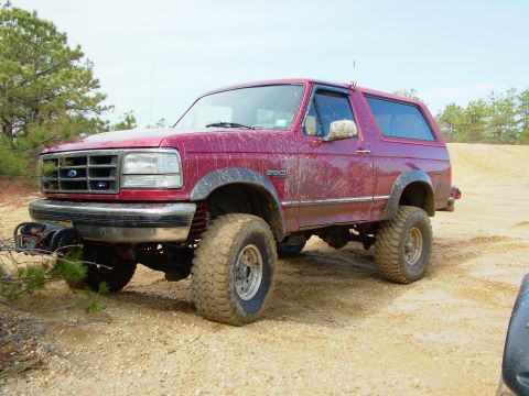 1994 Ford Bronco - A muddy red 1992 Ford Bronco sits on a dirt path, its off-road tires and lifted suspension evident.