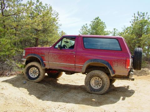 1994 Ford Bronco - A red Ford Bronco is shown from a three-quarter front angle on a dirt trail surrounded by trees.