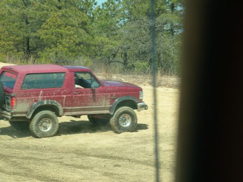1994 Ford Bronco - A red 1994 Ford Bronco is seen driving off-road on a dirt path surrounded by trees.