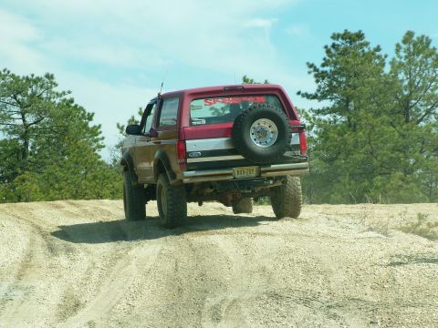 1994 Ford Bronco - Red Ford Bronco with a spare tire on the back drives on a dirt road under a blue sky.