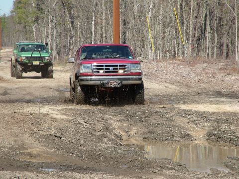 1994 Ford Bronco - A red 1994 Ford Bronco navigates a muddy trail, with a lifted green Jeep following closely behind.