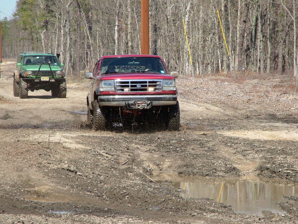 Red 1994 Ford Bronco driving through mud with a green Jeep behind it