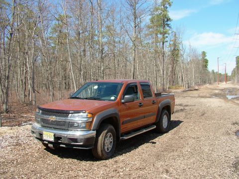 1994 Ford Bronco - An orange pickup truck is parked on a dirt road surrounded by trees.