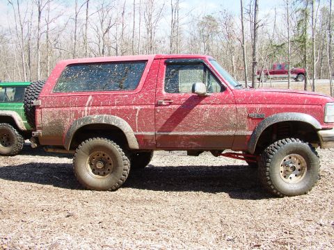 1994 Ford Bronco - Two off-road vehicles are parked on a dirt lot, with a third truck visible on a road in the distance.
