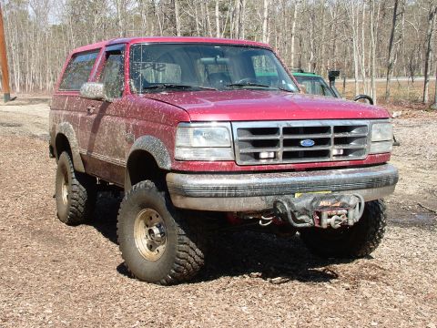 1994 Ford Bronco - A muddy red Ford Bronco is parked on a dirt road, with another vehicle visible behind it.