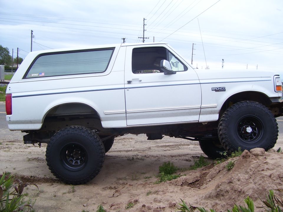 Lifted white full-size Ford Bronco flexing on a dirt mound, passenger side, mud tires and black wheels
