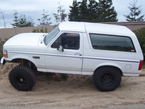 1994 Ford Bronco - A white Ford Bronco is shown lifted with large off-road tires, parked on a dirt surface with trees in the background.