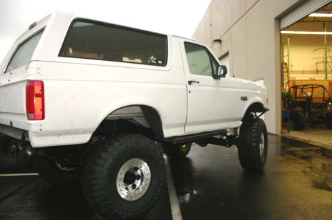 1995 Ford Bronco - A white 1995 Ford Bronco with aftermarket suspension and tires sits outside a garage, with another vehicle visible inside.